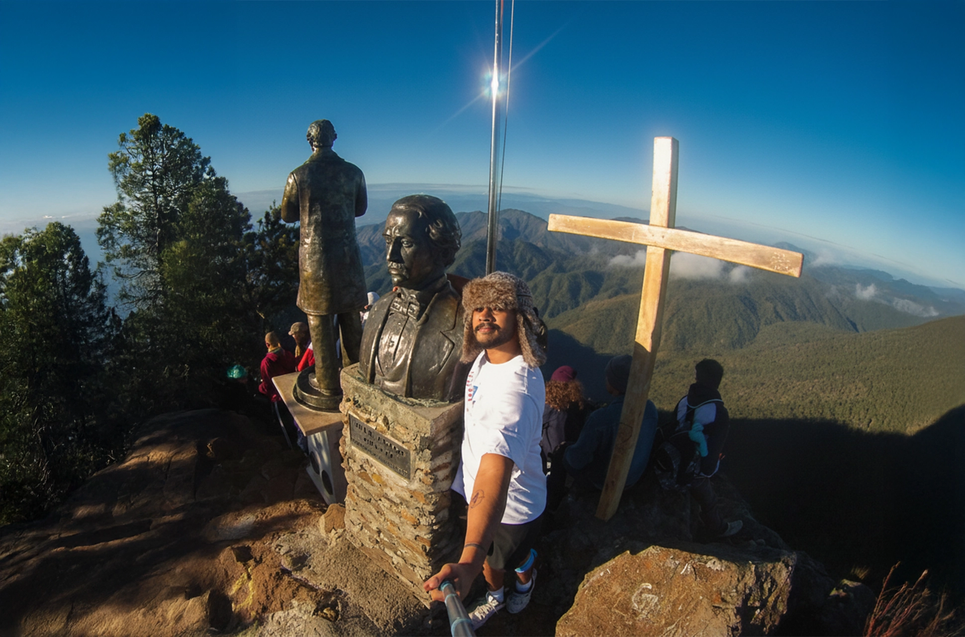 Group of hikers taking a selfie at Pico Duarte summit with the bust of Juan Pablo Duarte and a wooden cross