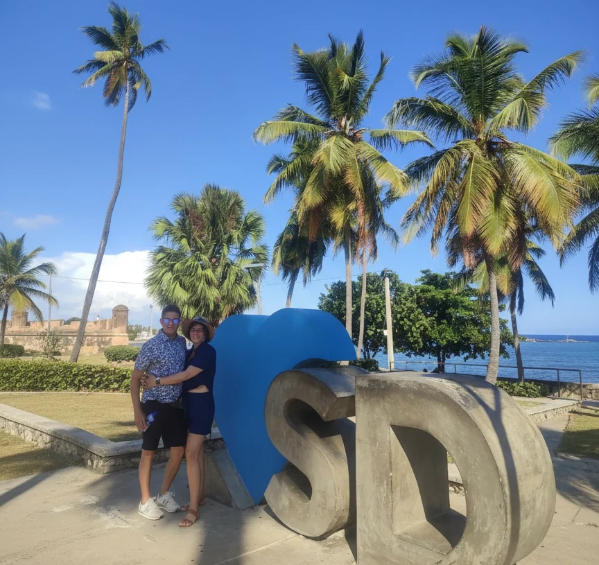 Couple posing at the SD sign with palm trees and the Caribbean Sea in Santo Domingo’s Malecón.